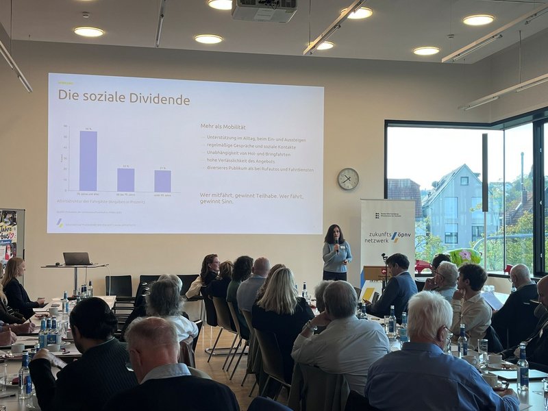 Vortragende steht vor Publikum in hellem Seminarraum mit Präsentation zum Thema soziale Dividende und Banner mit Logo 'zukunftsnetz ÖPNV'