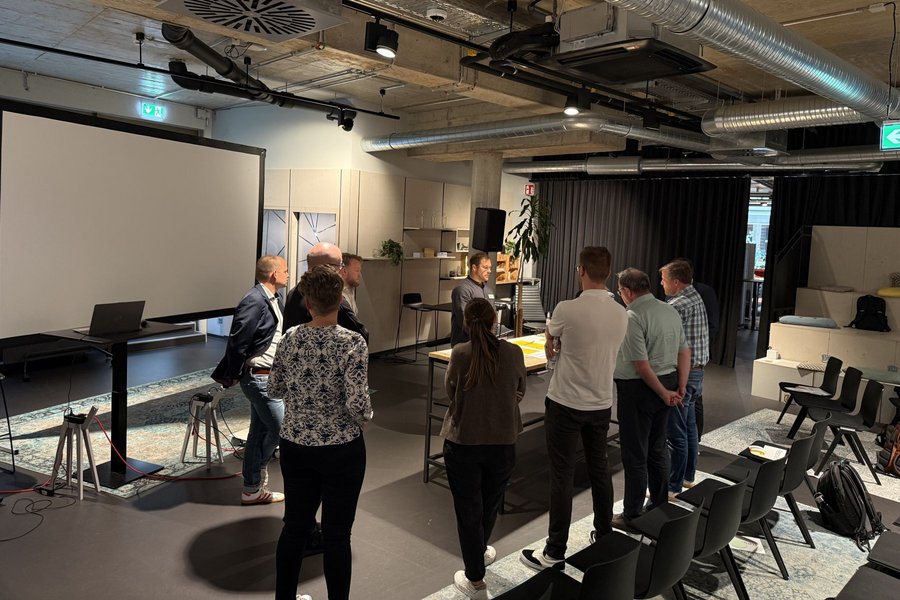Gruppe von Menschen steht in einem Raum mit Betonwänden und Decke um einem Tisch. Im Hintergrund hängt ein großen Bildschirm an der Wand.