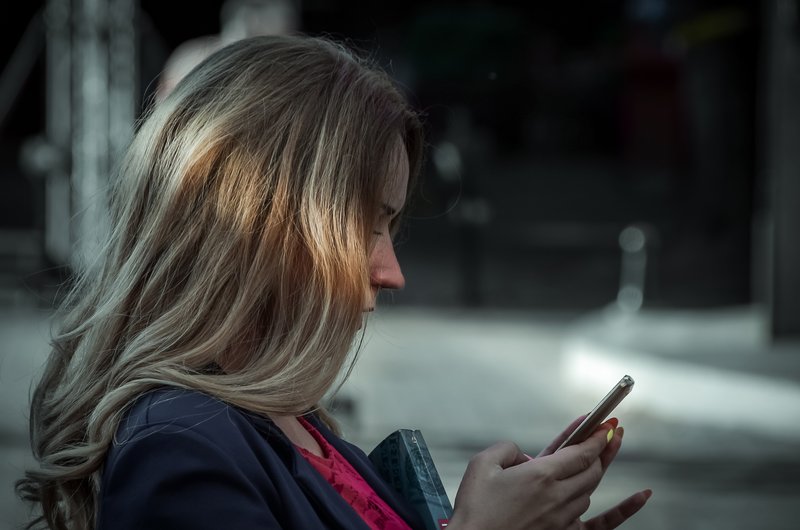 Blonde Frau sitzt und blickt auf das Smartphone in ihren Händen.