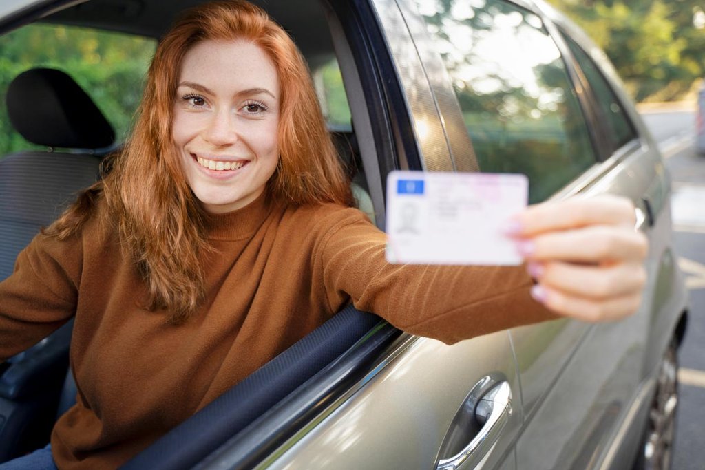 Eine junge Frau mit roten Haaren hält ihren Führerschein aus dem Autofenster.