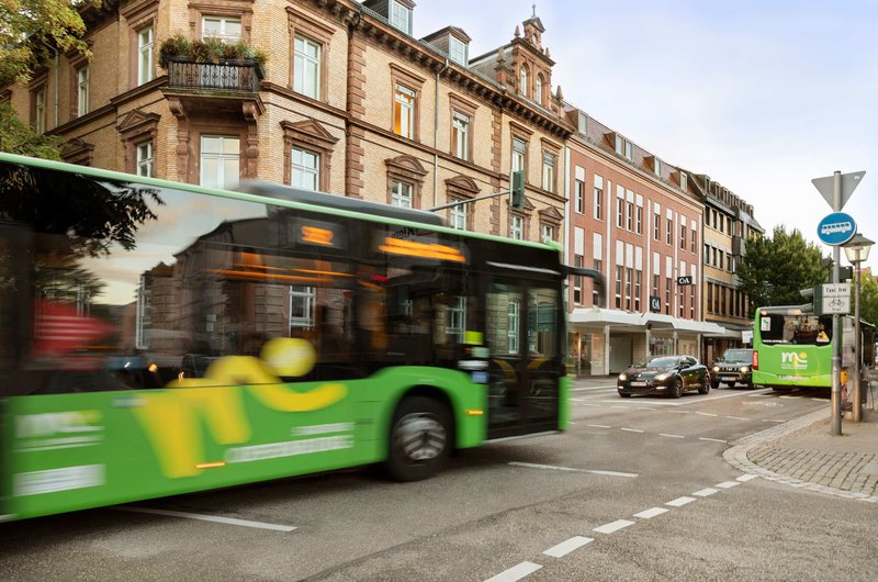 Grüner Linienbus fährt auf einer Busspur durch eine innerstädtische Kreuzung, umgeben von mehrstöckigen Gebäuden und weiterem Straßenverkehr.