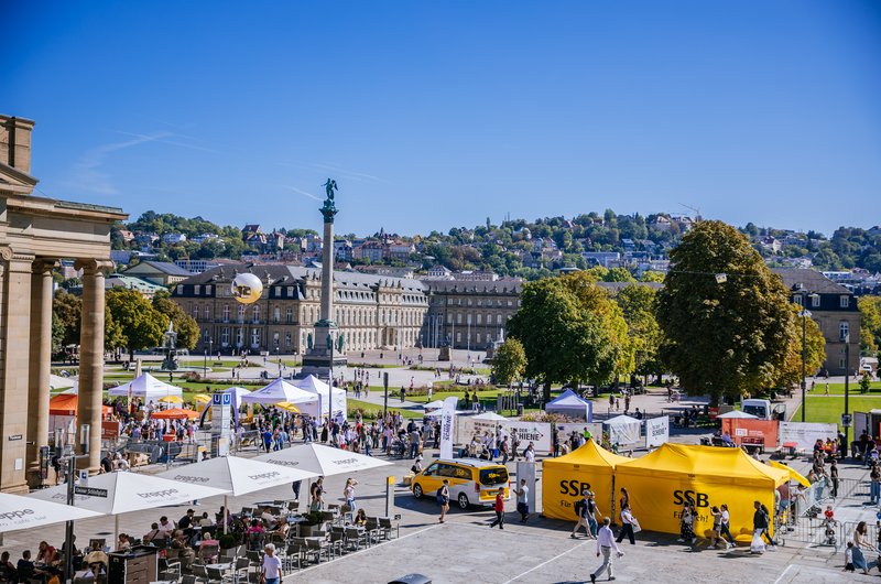 Blick über einen belebten Schlossplatz in Stuttgart mit Informationsständen, gelben SSB-Zelten, zahlreichen Besucherinnen und Besuchern sowie dem Neuen Schloss und der Jubiläumssäule im Hintergrund bei sonnigem Wetter.