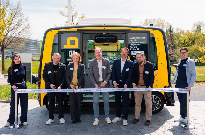 Sieben Personen stehen vor einem gelben autonomen Shuttle und halten ein Band mit dem Schriftzug 'Fraunhofer'