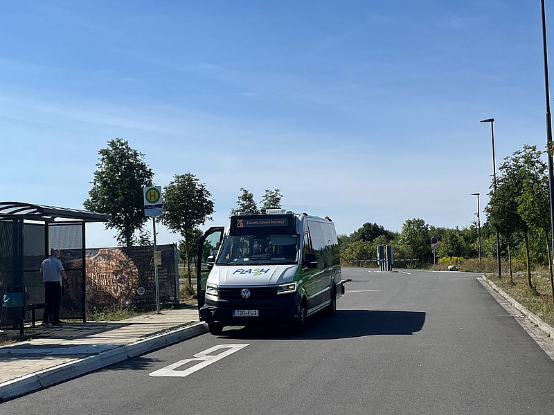 Kleinbus mit Aufschrift "FLASH" hält mit offener Tür auf Straße neben Bushaltestelle mit Wartehäuschen und zwei wartenden Personen