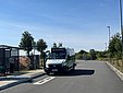 Kleinbus mit Aufschrift "FLASH" hält mit offener Tür auf Straße neben Bushaltestelle mit Wartehäuschen und zwei wartenden Personen