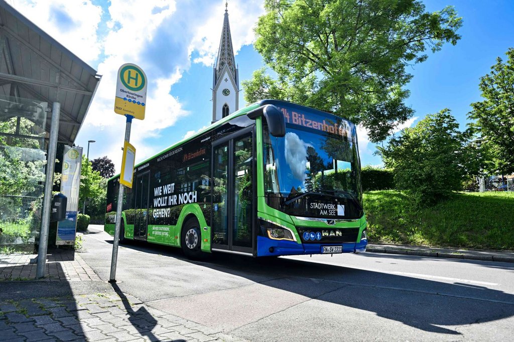 Grüner Stadtbus mit digitaler Linienanzeige vor Haltestellenschild an sonnigem Tag in einer Straße mit Bäumen und Kirche im Hintergrund