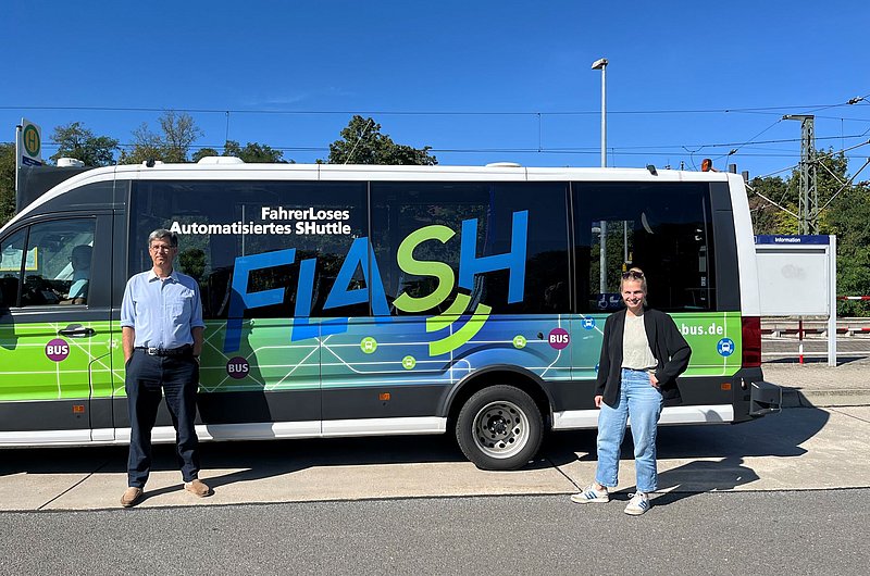 Zwei Personen stehen vor einem weißen Shuttlebus mit der Aufschrift 'Fahrerloses Automatisiertes SHuttle FLASH' und einem grünen und blauen Design