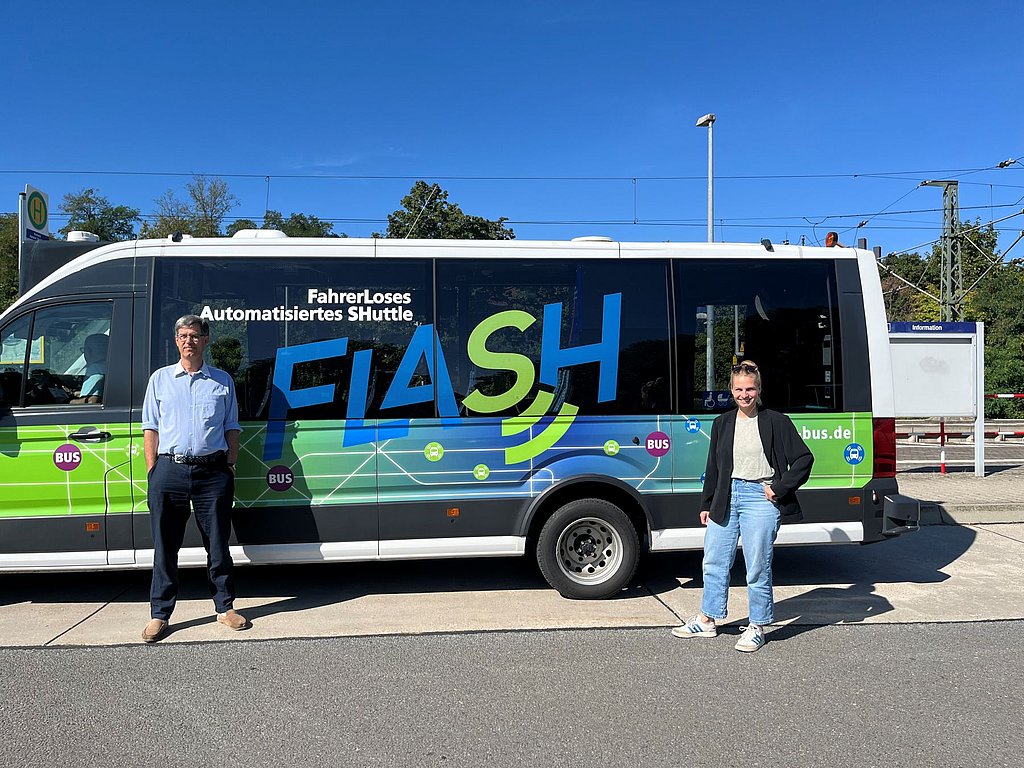 Zwei Personen stehen vor einem weißen Shuttlebus mit der Aufschrift 'Fahrerloses Automatisiertes SHuttle FLASH' und einem grünen und blauen Design