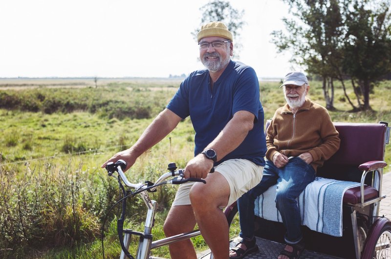 Ein Mann mit Mütze und Brille fährt einen älteren lächelnden Mann in einer Fahrradrikscha durch eine grüne Landschaft