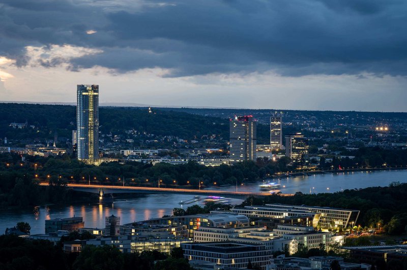 Abendliche Stadtansicht von Bonn am Rhein mit beleuchteten Hochhäusern, einer Brücke über den Fluss und dunklen Gewitterwolken am Himmel.