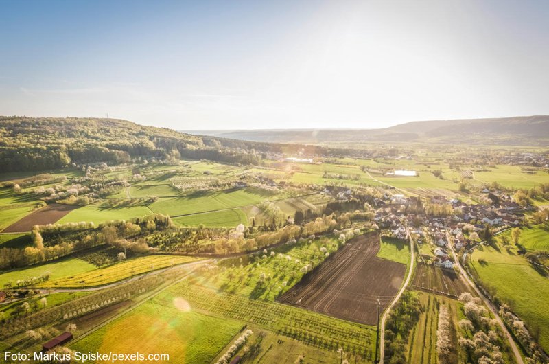 Grüne Landschaft mit einem Dorf und Zufahrtsstraßen die durch Felder verlaufen. Im Hintergrund sind bewaldete Hügel zu sehen.