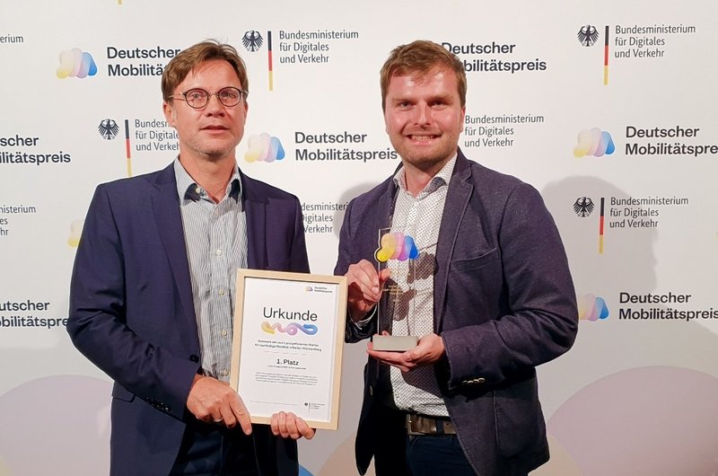 Christoph Erdmenger (Ministerium für Verkehr BW) und Sören Bröcker (NVBW) halten die Urkunde und die Trophäe hoch, die das Land Baden-Württemberg als Gewinner des deutschen Mobilitätspreises erhalten hat.