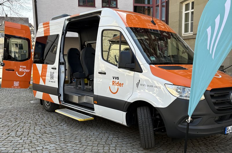 Ein weiß-orangener Kleinbus mit der Aufschrift VVS-Rider Rems-Murr-Kreis vor einem großen Gebäude; der rechte Teil ist ein Fachwerkhaus. 