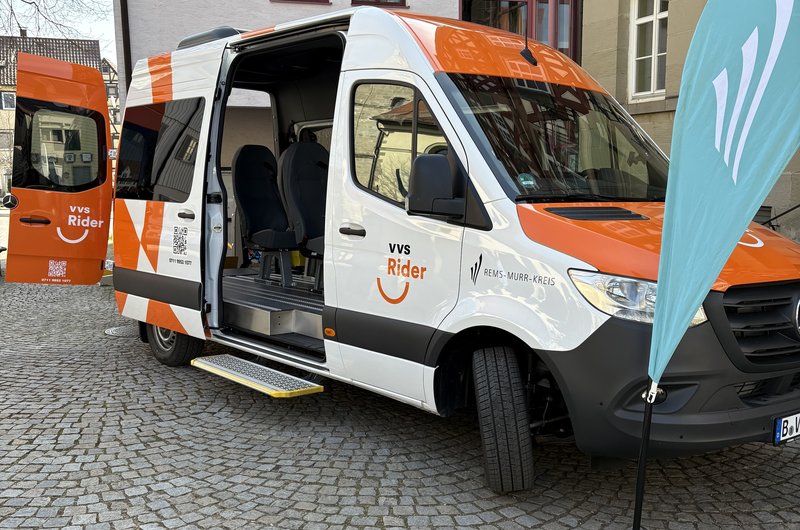 Ein weiß-orangener Kleinbus mit der Aufschrift VVS-Rider Rems-Murr-Kreis vor einem großen Gebäude; der rechte Teil ist ein Fachwerkhaus.