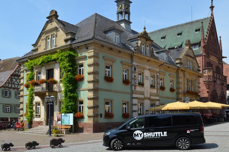 Ein schwarzer Kleinbus mit dem Aufdruck MyShuttle steht vor dem historischen grünen Rathaus in Bretten.