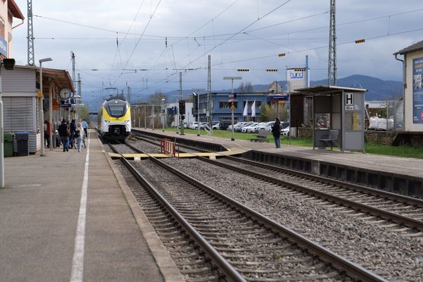 Bahnsteig mit ankommendem Zug, wartenden Personen und Fahrgastunterstand an einem Bahnhof mit Bergen im Hintergrund
