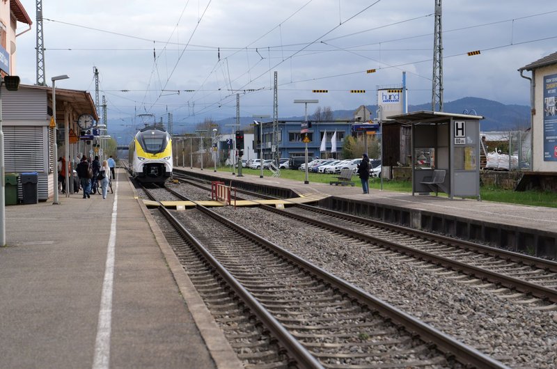 Bahnsteig mit ankommendem Zug, wartenden Personen und Fahrgastunterstand an einem Bahnhof mit Bergen im Hintergrund