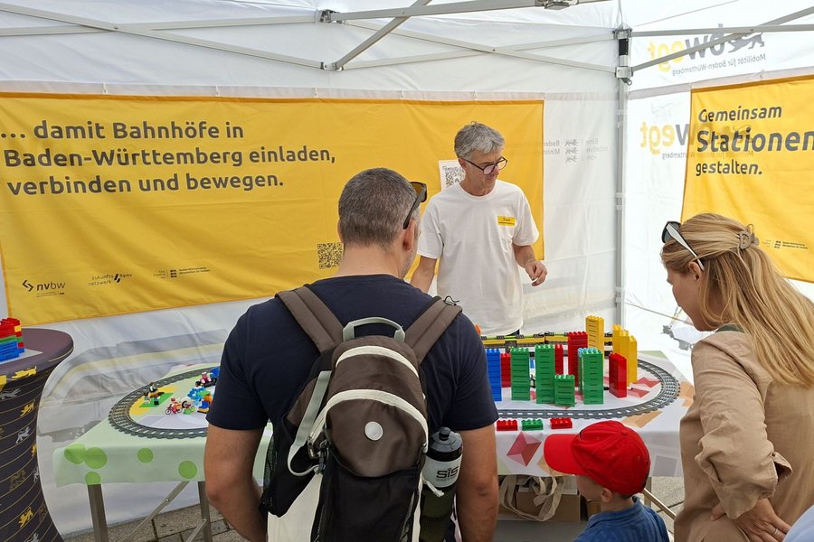 Personen stehen an einem Stand mit einer Modelleisenbahn und einem gelben Banner mit Text zu Bahnhöfen in Baden-Württemberg