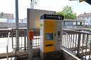 Fahrkartenautomat mit gelbem und grauem Gehäuse auf einem Bahnsteig vor Gleisen und Bahnhofsgebäude