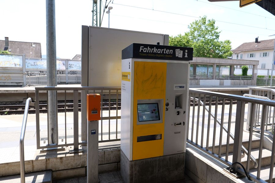 Fahrkartenautomat mit gelbem und grauem Gehäuse auf einem Bahnsteig vor Gleisen und Bahnhofsgebäude