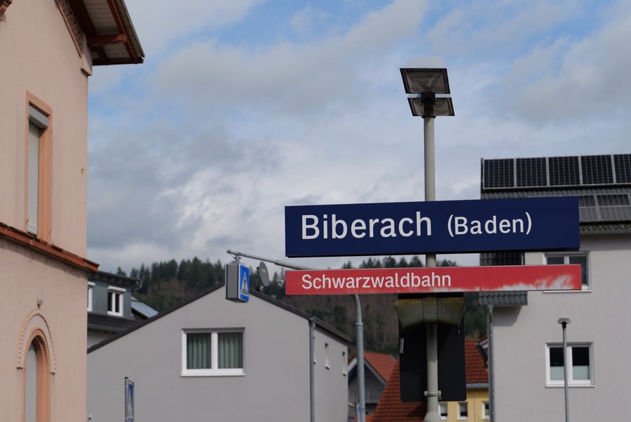Bahnhofsschild mit der Aufschrift 'Biberach (Baden)' und darunter ein rotes Schild mit der Aufschrift 'Schwarzwaldbahn' vor Häusern und bewaldetem Hügel im Hintergrund