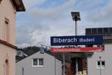Bahnhofsschild mit der Aufschrift 'Biberach (Baden)' und darunter ein rotes Schild mit der Aufschrift 'Schwarzwaldbahn' vor Häusern und bewaldetem Hügel im Hintergrund