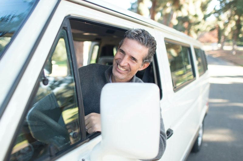 Ein Mann mittleren Alters sitzt in einem weißen Kleinbus und er streckt seinen Kopf auf dem Fahrerfenster. Dabei sieht man ihn aus einer schrägen Vorderansicht.Auto befindet sich auf einer Landstraße.