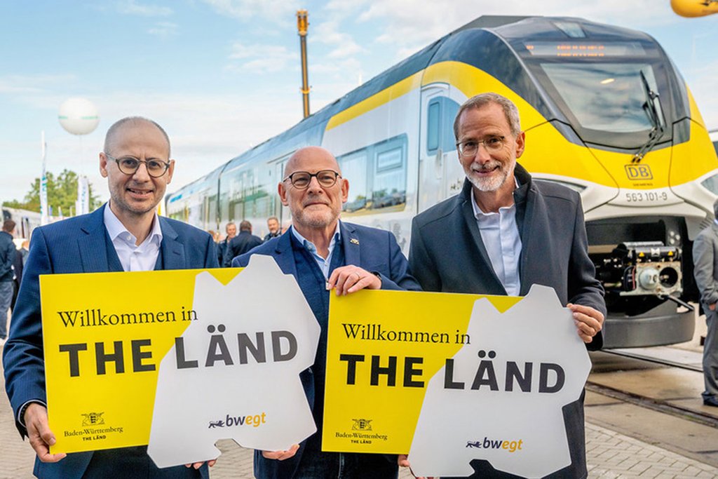 Drei Männer stehen in einer Reihe und halten Pappen hoch, auf denen The Länd steht. Der Man in der Mitte ist Baden-Württembergs Verkehrsminister Winfried Hermann. Die Männer befinden sich auf der Messe InnoTrans und stehen vor einem gelb-weißen Zug im bwegt-Design.