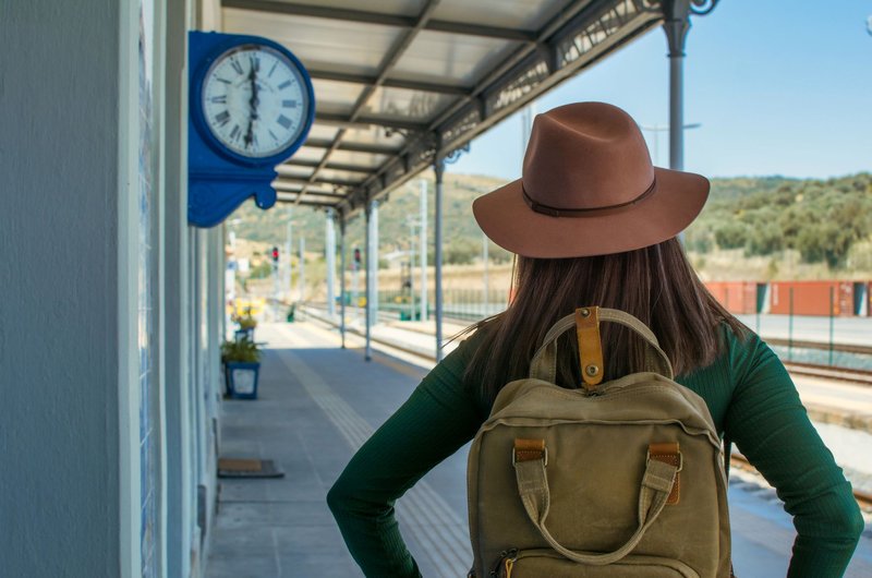 Frau mit braunem Hut und Rucksack steht am Bahnsteig und blickt auf eine Bahnhofsuhr.