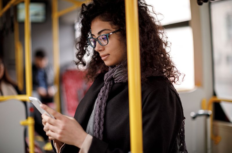 Eine Frau mit lockigen schwarzen Haaren und Brille steht in einem Bus und schaut auf ihr Smartphone.