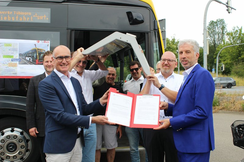 Mehrere Männer stehen vor einem Bus, zwei von ihnen halten gemeinsam einen roten Ordner mit Dokumenten.