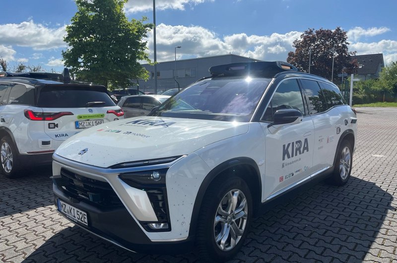Weißer SUV mit der Aufschrift 'KIRA' auf der Seite steht auf gepflastertem Parkplatz bei sonnigem Himmel