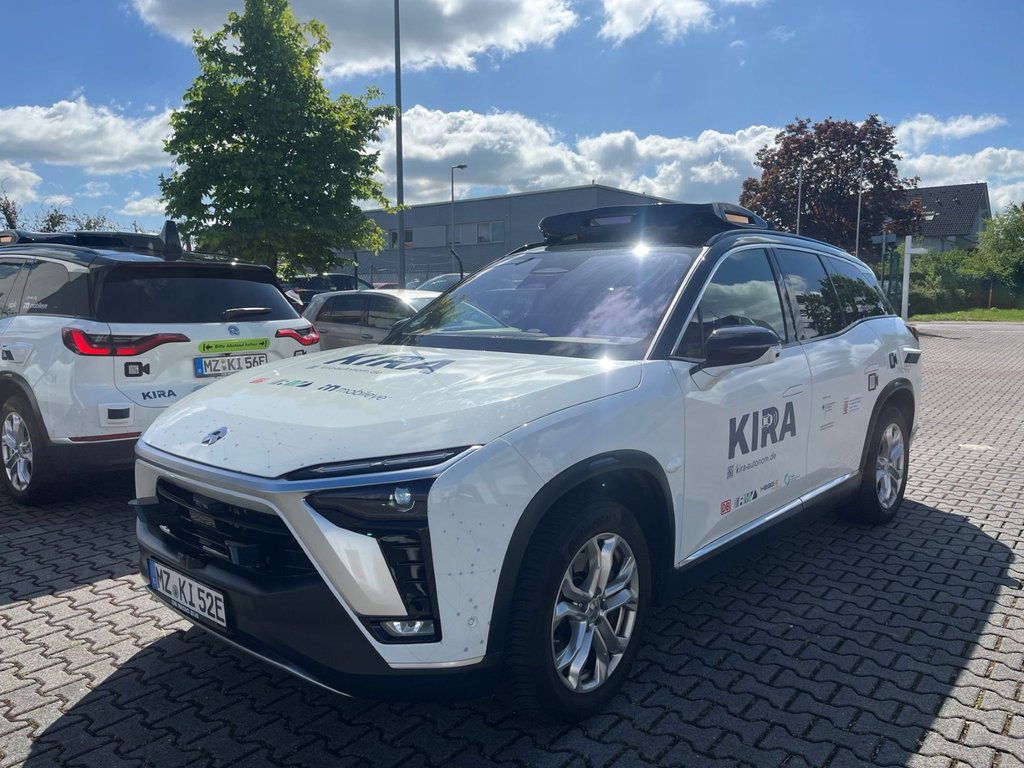 Weißer SUV mit der Aufschrift 'KIRA' auf der Seite steht auf gepflastertem Parkplatz bei sonnigem Himmel