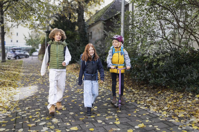 Drei Schulkinder mit Schulrucksäcken sind auf einem von Laub bedeckten Gehweg unterwegs. Ein Kind fährt mit einem Roller, die beiden anderen gehen zu Fuß.
